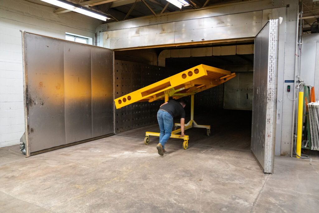Technician moving a freshly powder-coated yellow metal component into a curing oven at Biltwood Powder Coating.