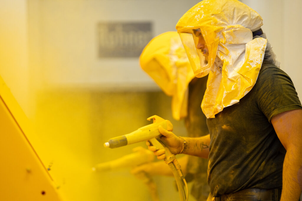 Powder coating technicians applying a yellow finish in a controlled spray booth at Biltwood Powder Coating.