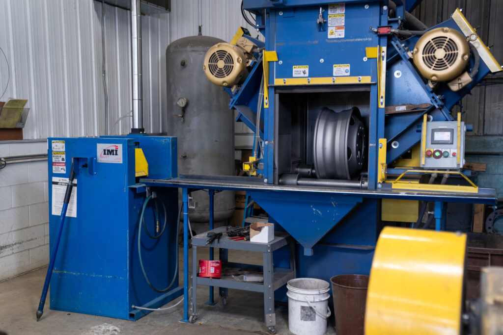 Industrial shot blasting machine at Biltwood Powder Coating processing a metal wheel for surface preparation.